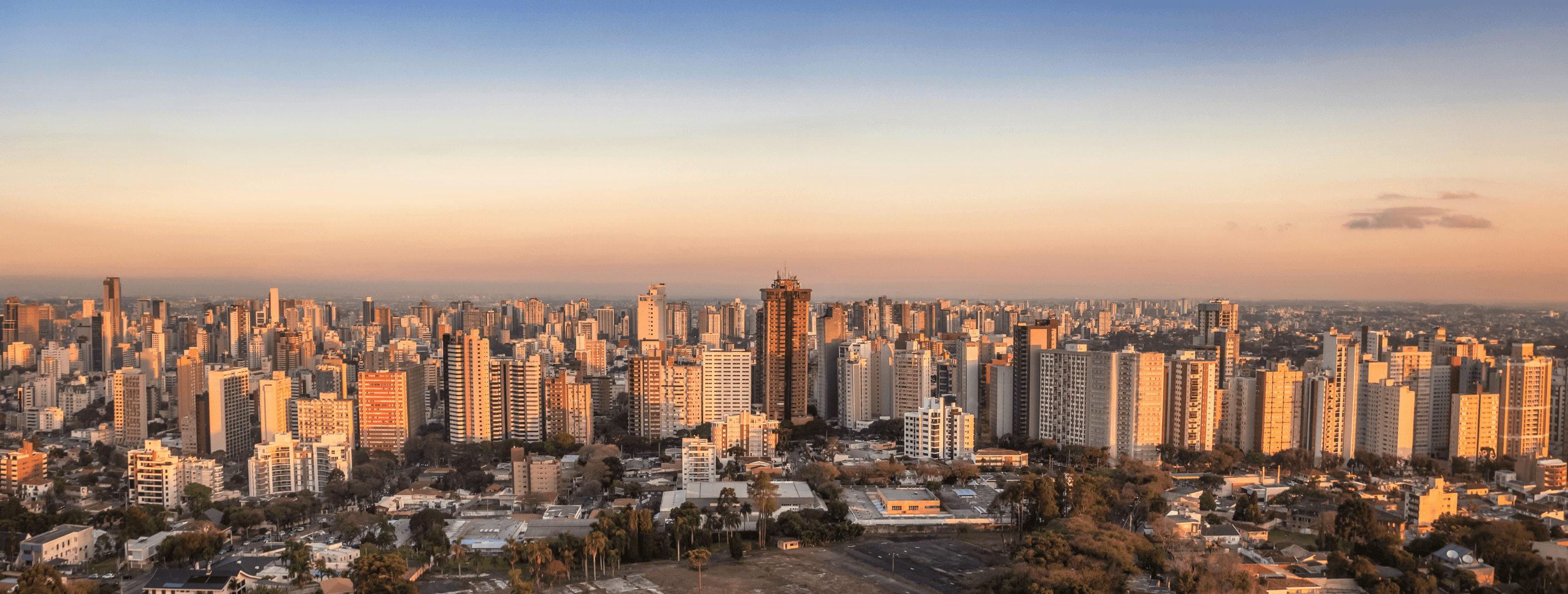Imagem de drone do bairro Mercês, em Curitiba, mostrando prédios altos, casas e o cenário urbano iluminado pelo sol do final da tarde.